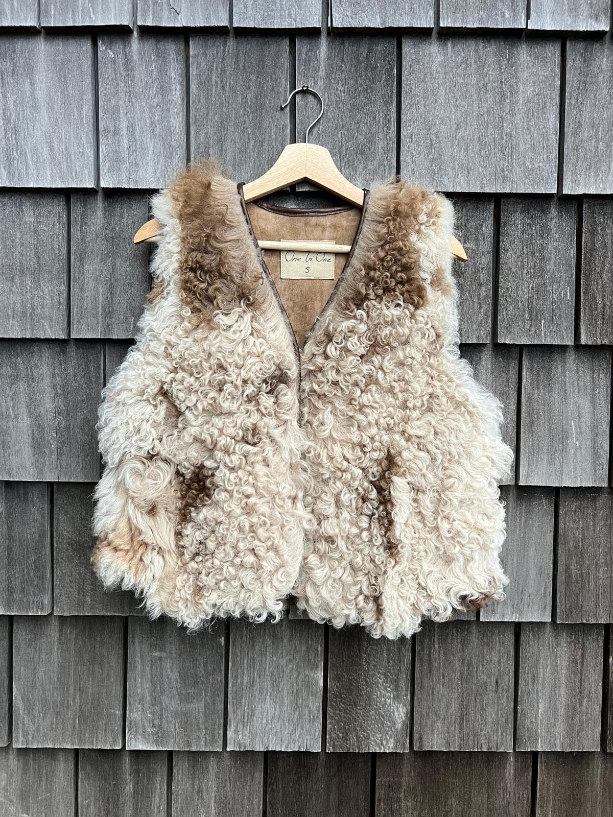 Handmade cream curly-wool sheepskin vest with caramel tips, on a wooden hanger against grey timber shingles