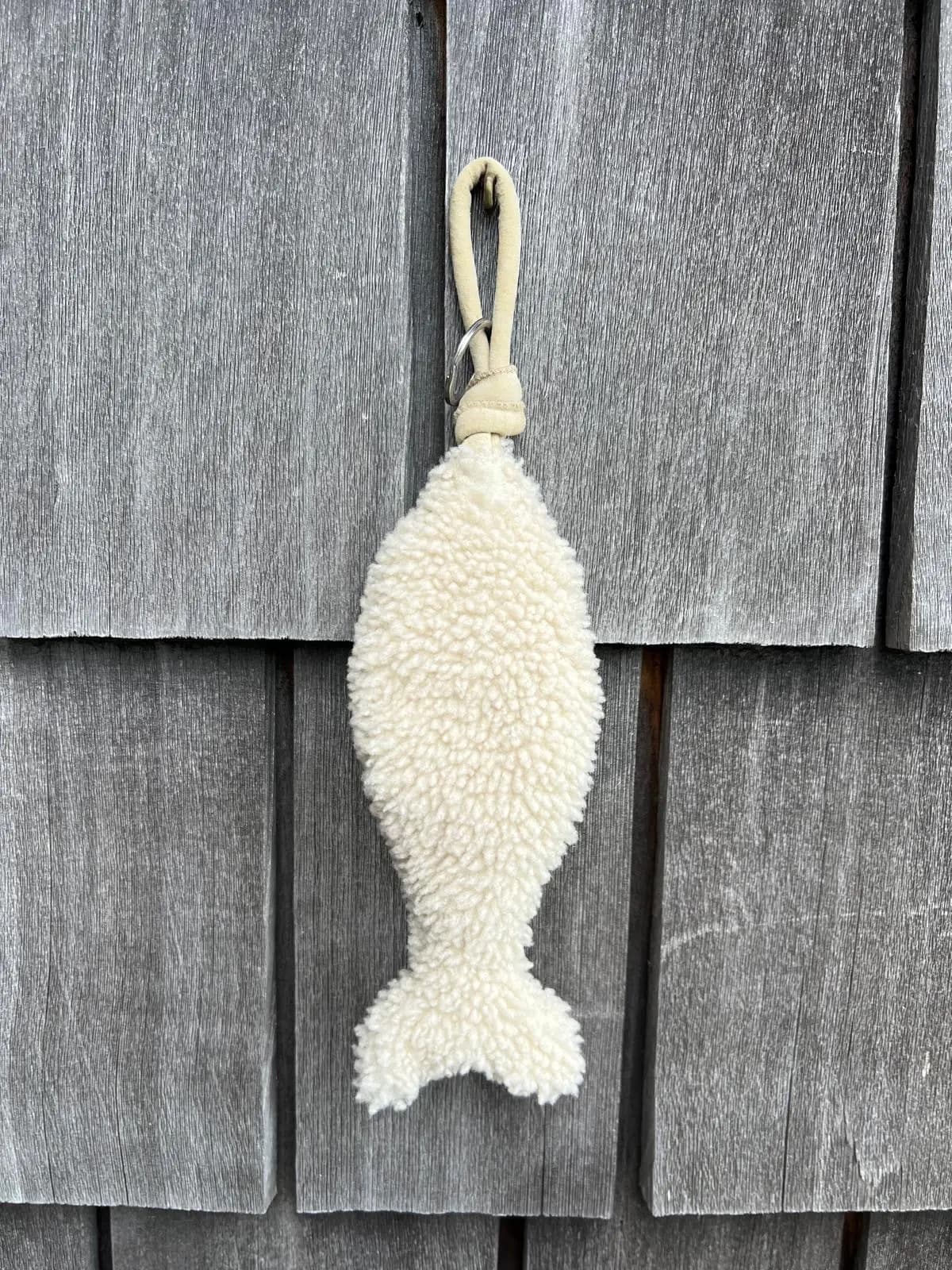 Handmade cream curly-wool sheepskin fish keychain with a suede loop and metal key ring, hanging against grey timber shingles