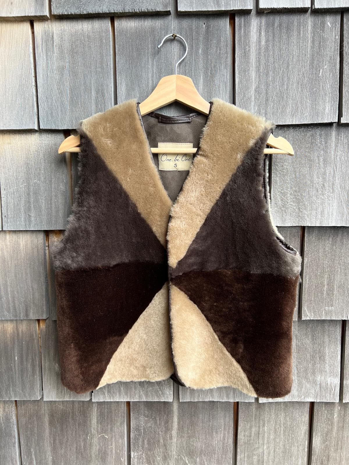 Front of a handmade patchwork shearling vest in taupe, grey-brown and deep brown panels with a hand-lettered One Bi One size S label, on a wooden hanger